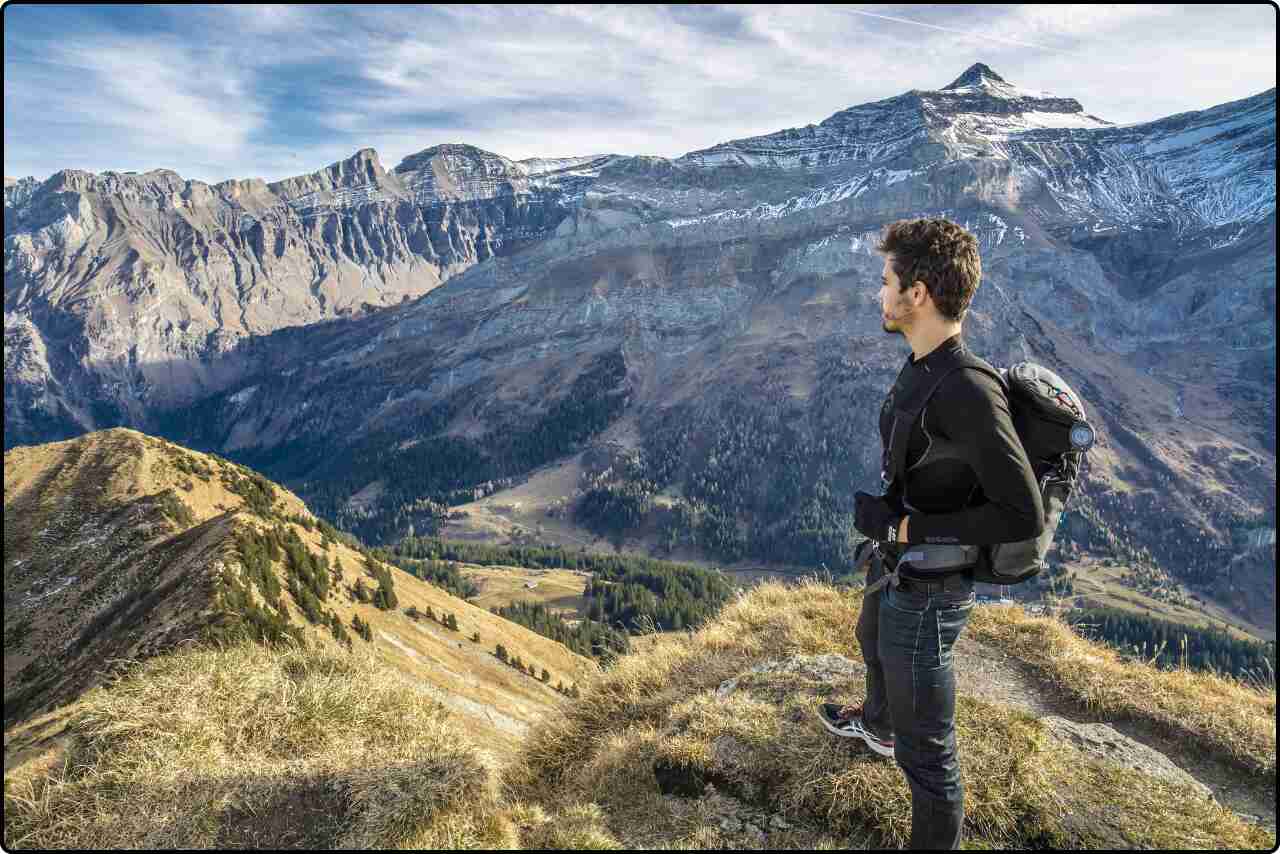 Man standing on top of a mountain, holding a trekking bag, surrounded by stunning scenery